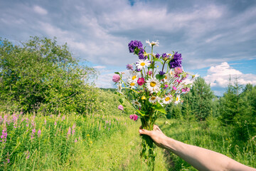 Fototapeta na wymiar A bouquet of wild flowers in your hand on the background of the river and the sky with clouds.