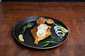 pomfret fish fry- deeply fried two pomfret fishes which is well garnished with lemon and onion in a white ceramic plate with wooden background