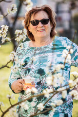 Smiling senior woman drinking coffee in blooming garden. Relaxation and enjoyment outdoors at springtime