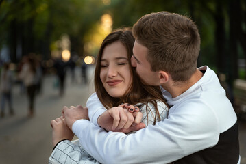 Portrait of happy couple in love in park. Guy hugs his beloved by shoulders and kiss