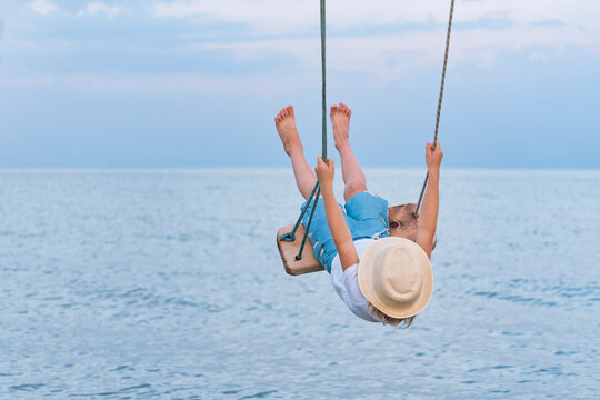 Child riding on rope swing over the water and lifted high legs. Vacation at sea