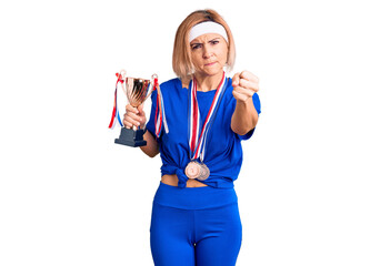 Young blonde woman holding champion trophy wearing medals annoyed and frustrated shouting with anger, yelling crazy with anger and hand raised