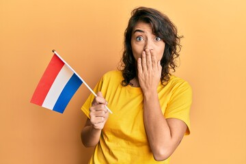 Young hispanic woman holding holland flag covering mouth with hand, shocked and afraid for mistake....