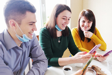 Friends with masks on their faces chat in a cafe and use mobile phones to view content on social networks - Concept of a new noram during the coronavirus epidemic - Positive young people