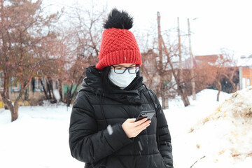 Fototapeta na wymiar A girl with a medical mask on her face on the street in winter during a snowfall with a smartphone in her hands. Mask mode.