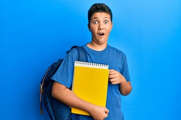 Teenager hispanic boy wearing school bag and holding books scared and amazed with open mouth for surprise, disbelief face