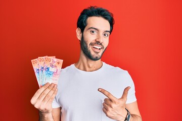 Young hispanic man holding swiss franc banknotes smiling happy pointing with hand and finger