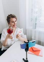 A woman who has a video call on her smartphone holds a glass of wine and shows a card with a heart. Remote acquaintance or declaration of love during quarantine. Virtual Valentine's Day.medium shot