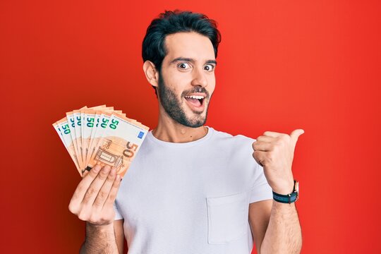 Young hispanic man holding euro banknotes pointing thumb up to the side smiling happy with open mouth