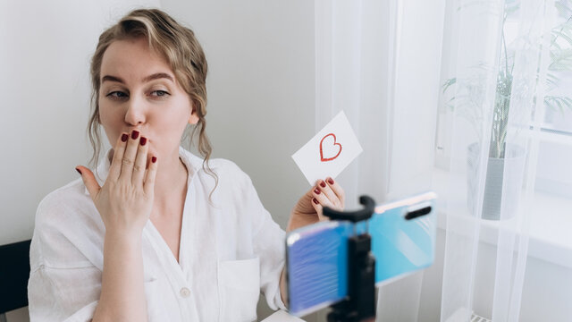 A woman who has a video call on her smartphone shows a drawing of a heart and blows a kiss. Remote acquaintance or declaration of love during quarantine. Virtual Valentine's Day.close-up