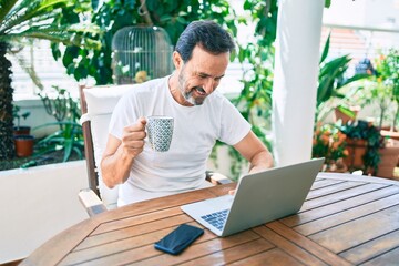 Middle age man with beard smiling happy at the terrace working from home using laptop drinking a cup of coffee