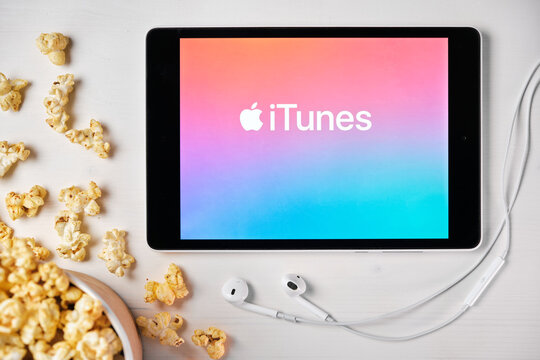 iTunes logo on the screen of the tablet laying on the white table and sprinkled popcorn on it. Apple earphones near the tablet, August 2020, San Francisco, USA