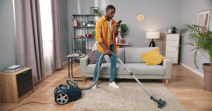 Handsome joyful African American young guy vacuuming modern living room and smiling typing on smartphone, male working cleans house using vacuum, man housekeeper, cleaning concept