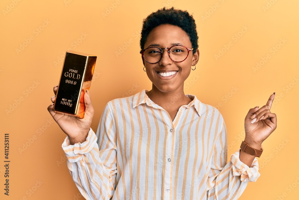 Poster young african american girl holding gold ingot smiling happy pointing with hand and finger to the si