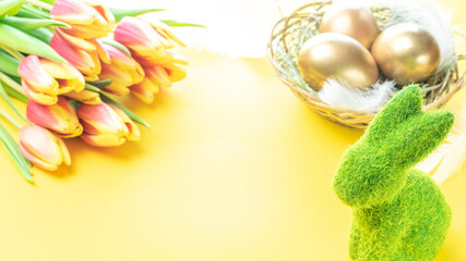 Easter Golden eggs in basket with spring tulips, white feathers on pastel yellow background in Happy Easter decoration. Spring holiday concept.
