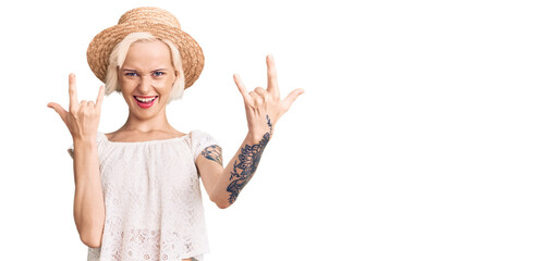 Young blonde woman with tattoo wearing summer hat shouting with crazy expression doing rock symbol...
