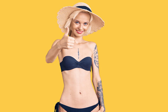 Young blonde woman with tattoo wearing bikini and summer hat doing happy thumbs up gesture with hand. approving expression looking at the camera showing success.