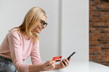 A smiling middle-aged woman holds a credit card and a smartphone indoor, going to pay online order, get or send payment. Contented charming mature lady enjoying online shopping at home