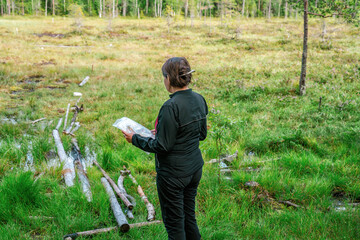 Cute middle aged Caucasian women wearing black sportswear standing at marsh edge and looking at map during exercise in outdoor orienteering in pine tree forest, Sweden, hobby sport, side view