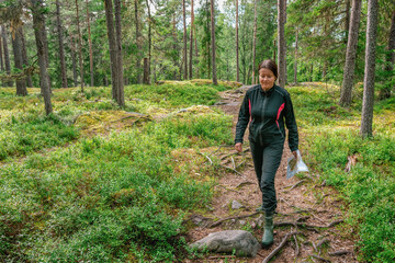 Cute middle aged Caucasian women wearing black sportswear walking with map in pine tree forest during exercise in outdoor orienteering, Sweden, hobby sport, front view