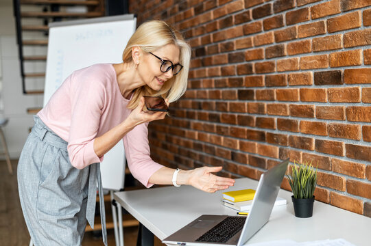 Modern And Skilled Middle-aged Business Woman Looks At The Laptop Screen And Recording A Voicemail On The Smartphone, Female Project Manager Makes A Remark And Sends To An Executor