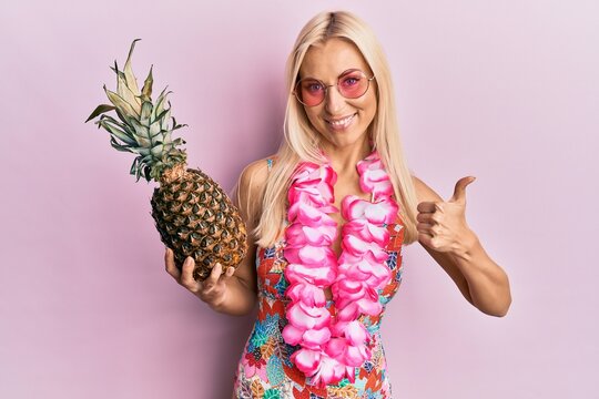 Young blonde woman wearing swimsuit and hawaiian lei holding pineapple smiling happy and positive, thumb up doing excellent and approval sign