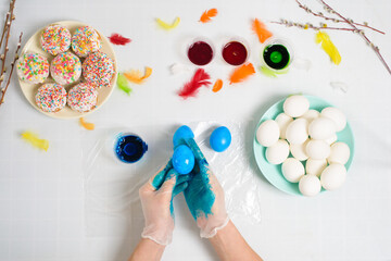 the process of coloring eggs for Easter in blue and red colors. a woman in gloves paints eggs with dye from a jar, top view, willow branches and colorful feathers decor