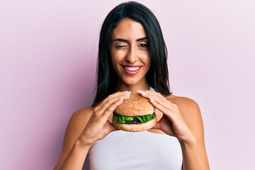 Beautiful hispanic woman eating a tasty classic burger winking looking at the camera with sexy expression, cheerful and happy face.