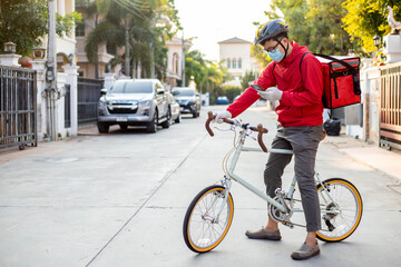 A courier on a bicycle with a red backpack wearing a mask and a helmet to deliver orders and packages for customers.
