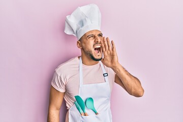 Young arab man wearing professional cook apron and hat shouting and screaming loud to side with hand on mouth. communication concept.