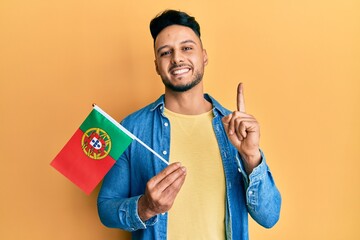 Young arab man holding portugal flag smiling with an idea or question pointing finger with happy face, number one