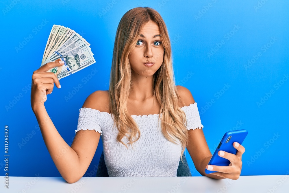 Poster beautiful blonde young woman holding dollars and using smarpthone puffing cheeks with funny face. mo