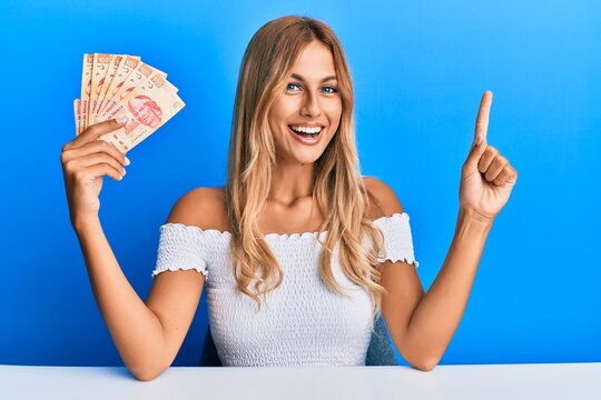 Beautiful blonde young woman holding 100 mexican pesos banknotes smiling with an idea or question pointing finger with happy face, number one