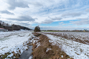 Verschneite Felder und gefrorerner Bach  mit Wolken und blauem Himmel im Winter