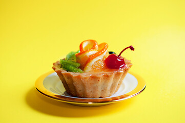 Juicy fruit tartlet cake on a white saucer with a golden border with orange, candied cherry and kiwi on a yellow background, top view with copy space. Confectionery, cooking