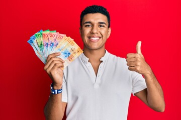 Young arab man holding swiss franc banknotes smiling happy and positive, thumb up doing excellent and approval sign