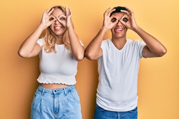Young interracial couple wearing casual white tshirt doing ok gesture like binoculars sticking tongue out, eyes looking through fingers. crazy expression.