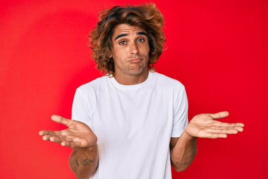 Young hispanic man wearing casual white tshirt clueless and confused expression with arms and hands raised. doubt concept.