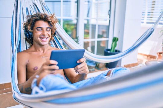 Young hispanic man relaxed using tablet and headphones lying on the hammock at terrace.