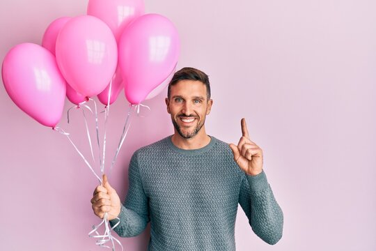 Handsome man with beard holding pink balloons surprised with an idea or question pointing finger with happy face, number one
