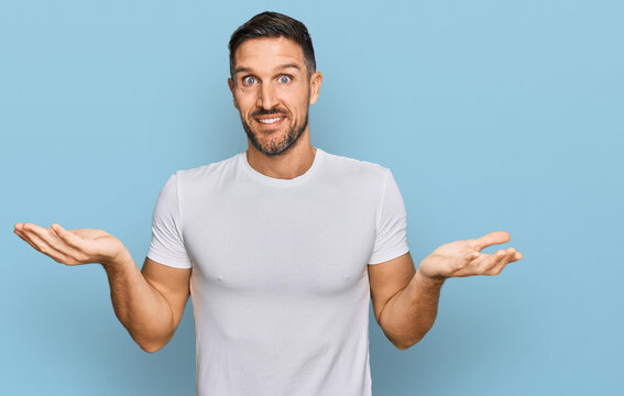 Handsome man with beard wearing casual white t shirt clueless and confused expression with arms and hands raised. doubt concept.