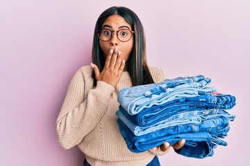 Young latin girl holding stack of folded jeans covering mouth with hand, shocked and afraid for...