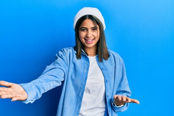 Young latin woman wearing cute wool cap smiling cheerful offering hands giving assistance and acceptance.