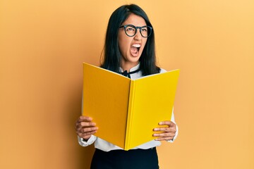 Beautiful asian young woman reading a book wearing glasses angry and mad screaming frustrated and furious, shouting with anger looking up.