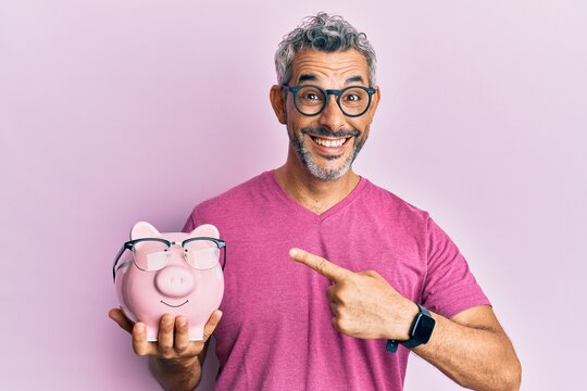 Middle age grey-haired man holding piggy bank with glasses smiling happy pointing with hand and finger