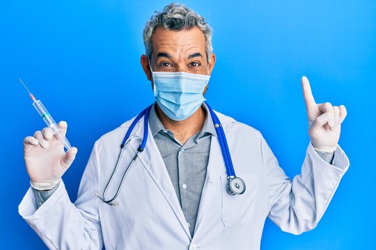 Middle age grey-haired man wearing doctor uniform and medical mask holding syringe smiling with an idea or question pointing finger with happy face, number one
