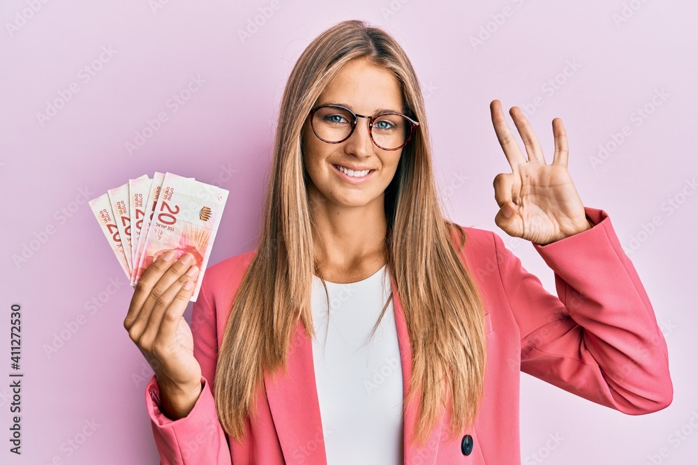 Wall mural Young blonde woman wearing business style holding israel shekels doing ok sign with fingers, smiling friendly gesturing excellent symbol