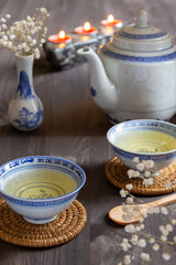 Top view of two cups of Chinese tea, blue teapot, vase, white flowers and candles, with selective focus, on dark table, in vertical