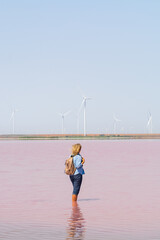 Woman standing on salt plains overlooking wind turbines. Water colored with pink algae.