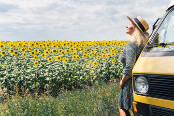 Young blonde woman travelling by campervan. Overlooking sunflower field on sunny summer day....